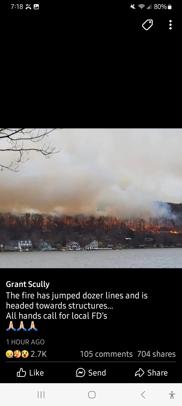 A screenshot of a Facebook post by Grant Scully showing a large wildfire burning across a hillside behind a lake, with…