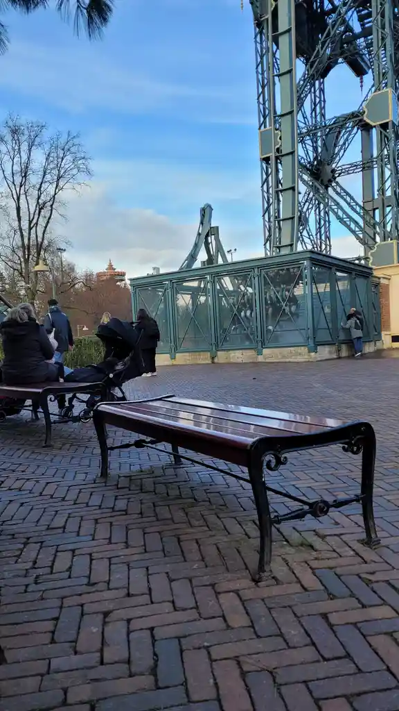 An empty wooden park bench with ornate black metal legs sits prominently in the foreground on a brick-paved area. Behind it…