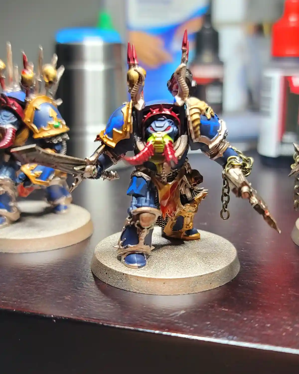 A close-up photo shows a heavily painted, ornate fantasy miniature figure in blue and gold armor, standing on a textured…