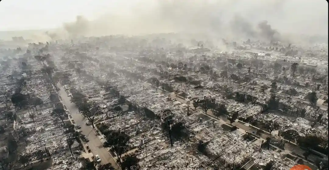 An aerial view shows a residential neighborhood completely destroyed by fire, with charred trees, rubble, and smoke rising…