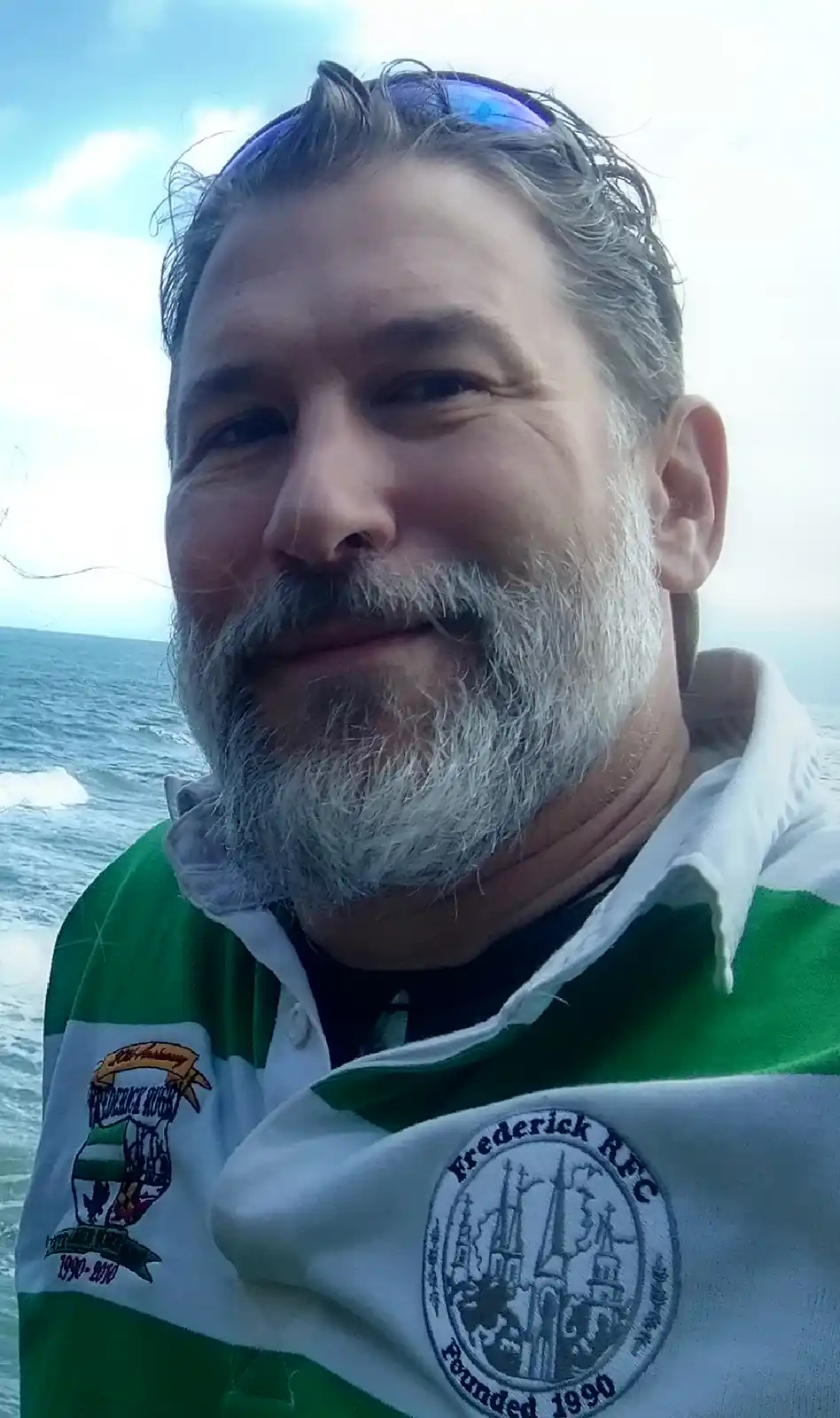 A middle-aged man with a gray beard and sunglasses on his head smiles at the camera while standing by the ocean, wearing a…