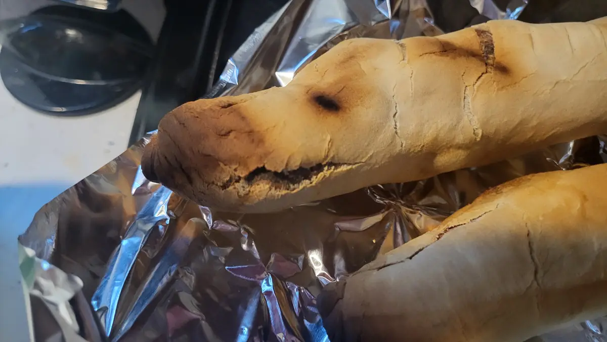 A close-up photo shows two long, crusty bread rolls resting on crumpled aluminum foil. One roll has a dark, burnt spot on…