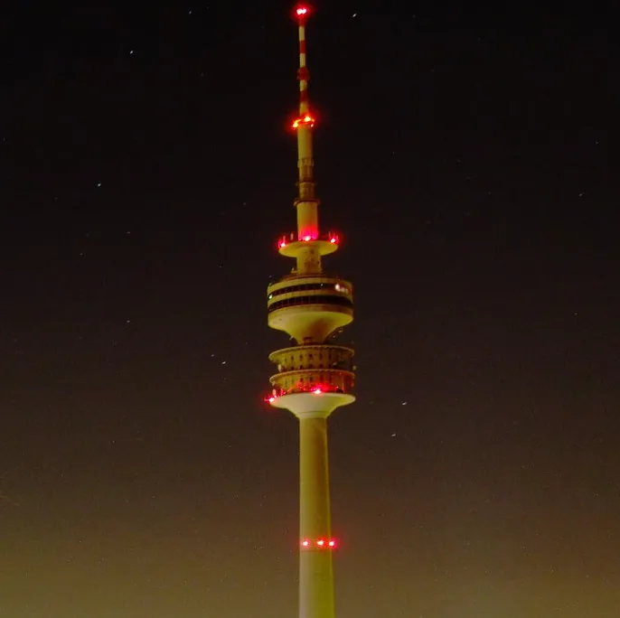 A tall, illuminated tower stands against a dark night sky with visible stars. Red lights are spaced along its structure…