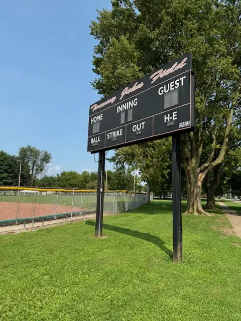 A black baseball scoreboard labeled 'Jimmy Johns Field' stands in a grassy field under a clear blue sky, with trees and a…