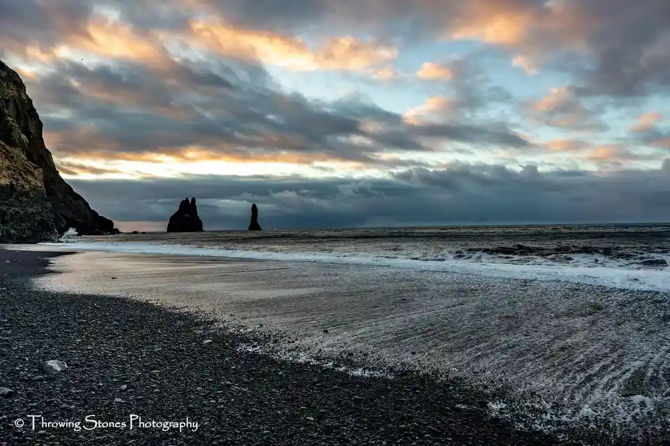A scenic photograph of a dark pebble beach at sunset or sunrise, with waves lapping the shore and dramatic clouds tinged…