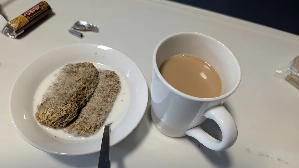 A white plate holds two dry, textured oatmeal bars soaking in milk, next to a white mug of tea. A partially unwrapped Rolo…