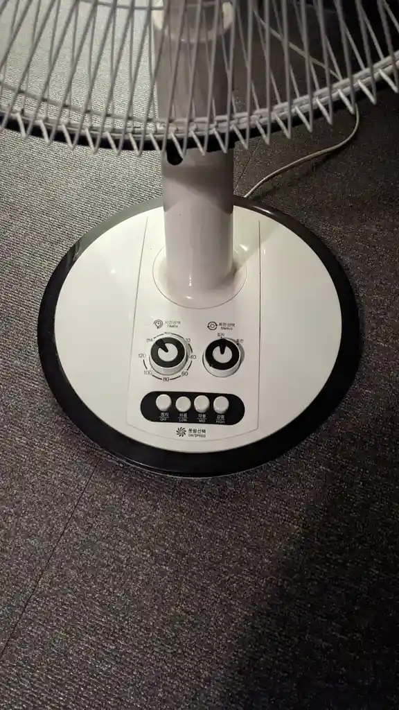 A close-up photo of a white pedestal fan's control panel on a dark speckled floor, showing a speed dial marked 40, 60, 80…