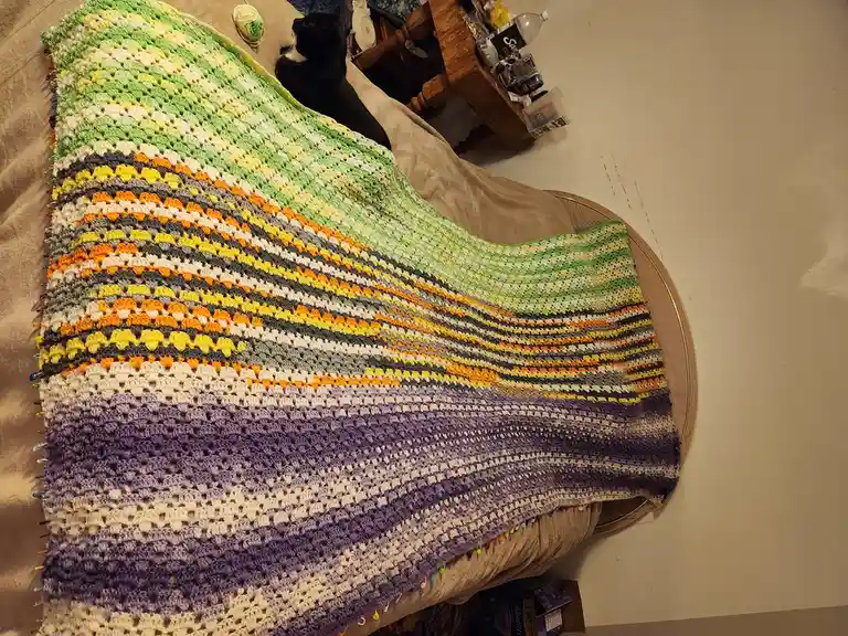 A large, colorful crocheted blanket with gradient stripes of green, yellow, orange, gray, and purple is draped over a bed or…