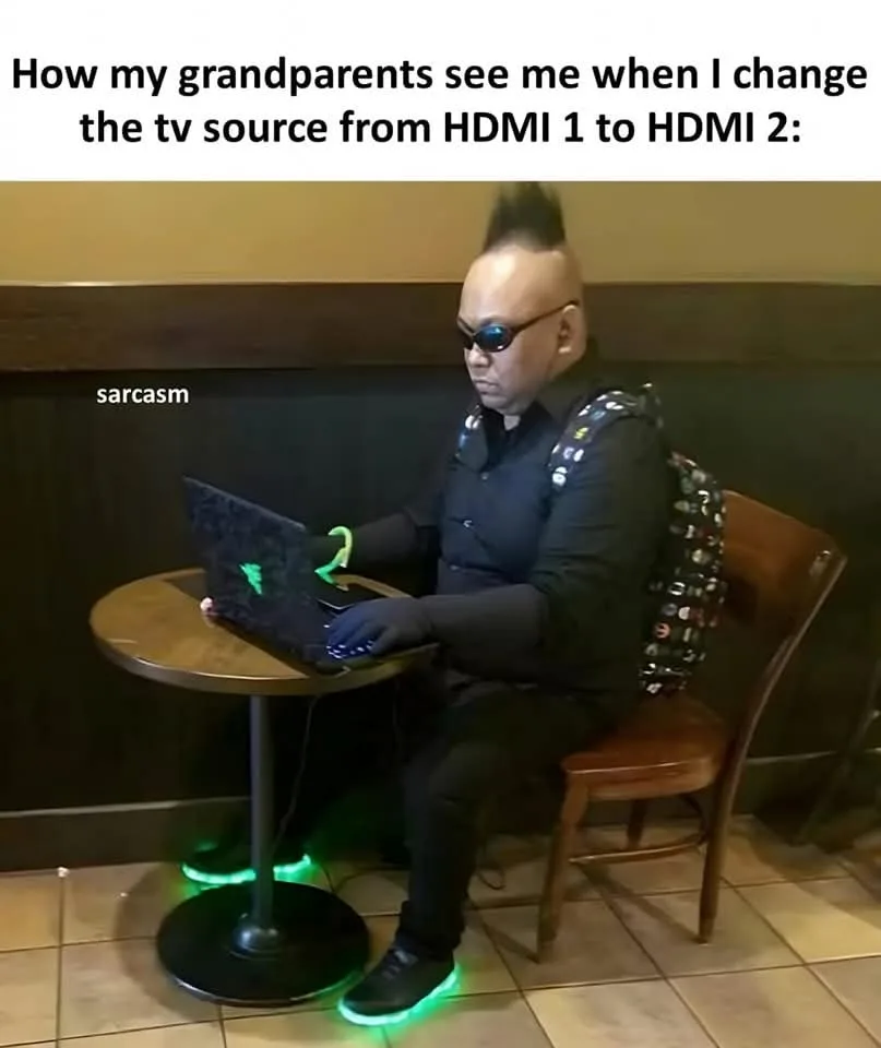 A man with a mohawk, sunglasses, and glowing green shoes sits at a table using a laptop. He wears a black outfit, a backpack…