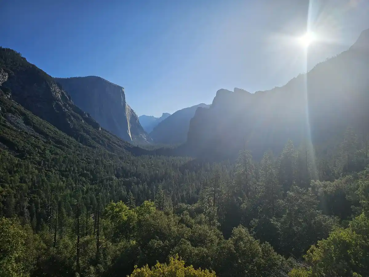 A scenic view of a mountain valley with dense forests and towering cliffs under a bright sun with lens flare. The overlaid…