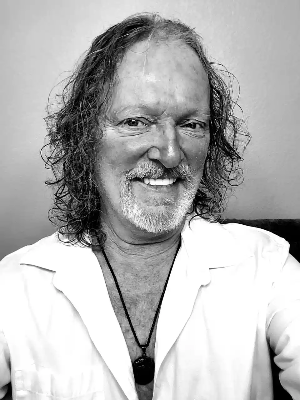 A black-and-white selfie of a smiling man with long, curly hair and a goatee, wearing a white shirt open at the collar and a…