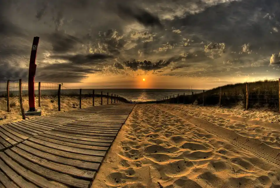 A dramatic, high-contrast beach sunset photo with a wooden boardwalk and sandy path leading to the ocean. The sky is filled…