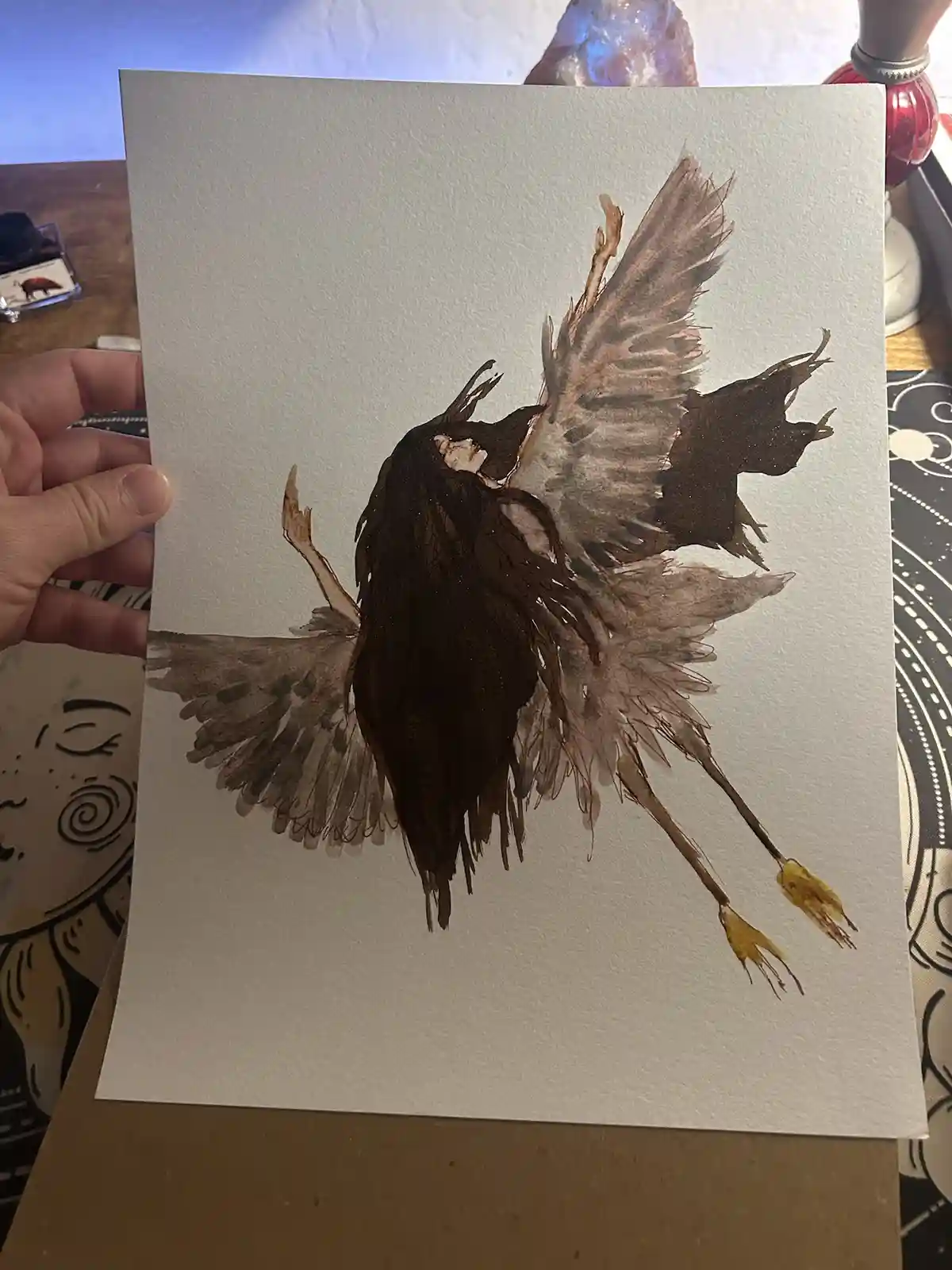 A hand holds up a watercolor painting of a dark, stylized bird with spread wings and yellow feet, rendered in brown and…