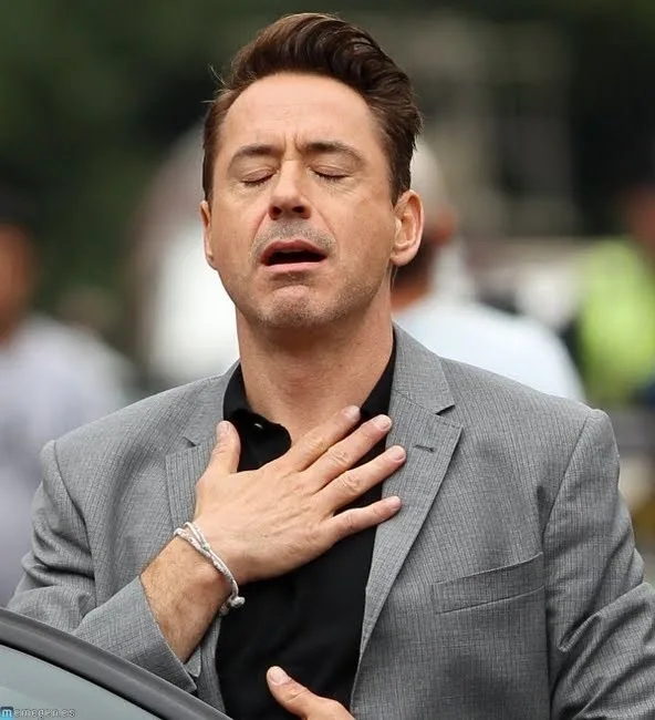 A man with dark hair and a gray suit has his eyes closed and mouth slightly open, placing his right hand over his heart in a…