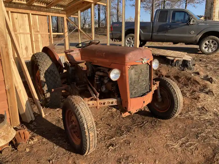 An old, rusty orange tractor sits in a dirt yard next to a partially built wooden shed, with a modern black pickup truck…