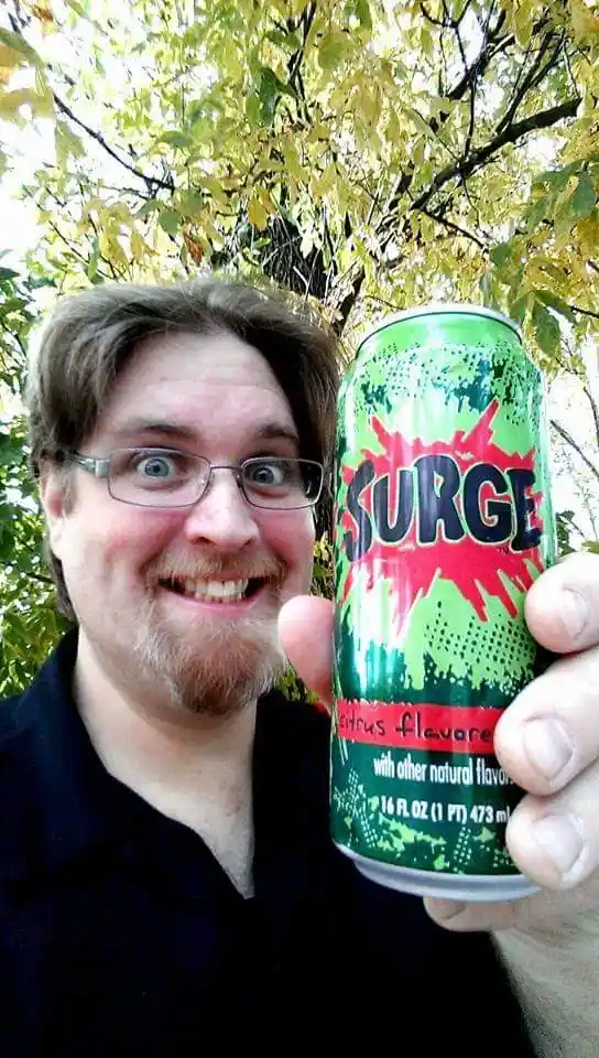 A man with glasses and a goatee holds up a green can of Surge citrus-flavored soda, his face contorted in an exaggerated…