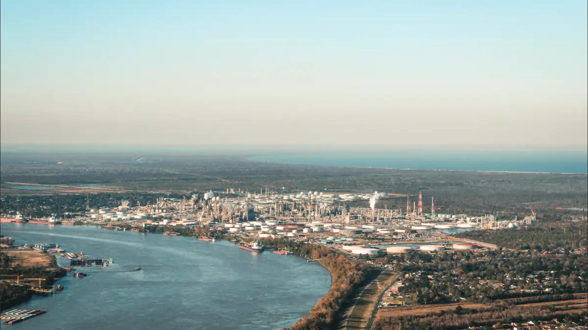 An aerial view of a large industrial complex with numerous storage tanks, pipes, and smokestacks along a riverbank, with a…