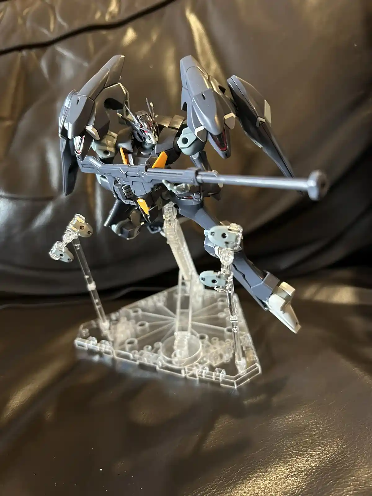 A gray and black Gundam model with orange accents is posed on a clear plastic display stand against a dark brown leather…
