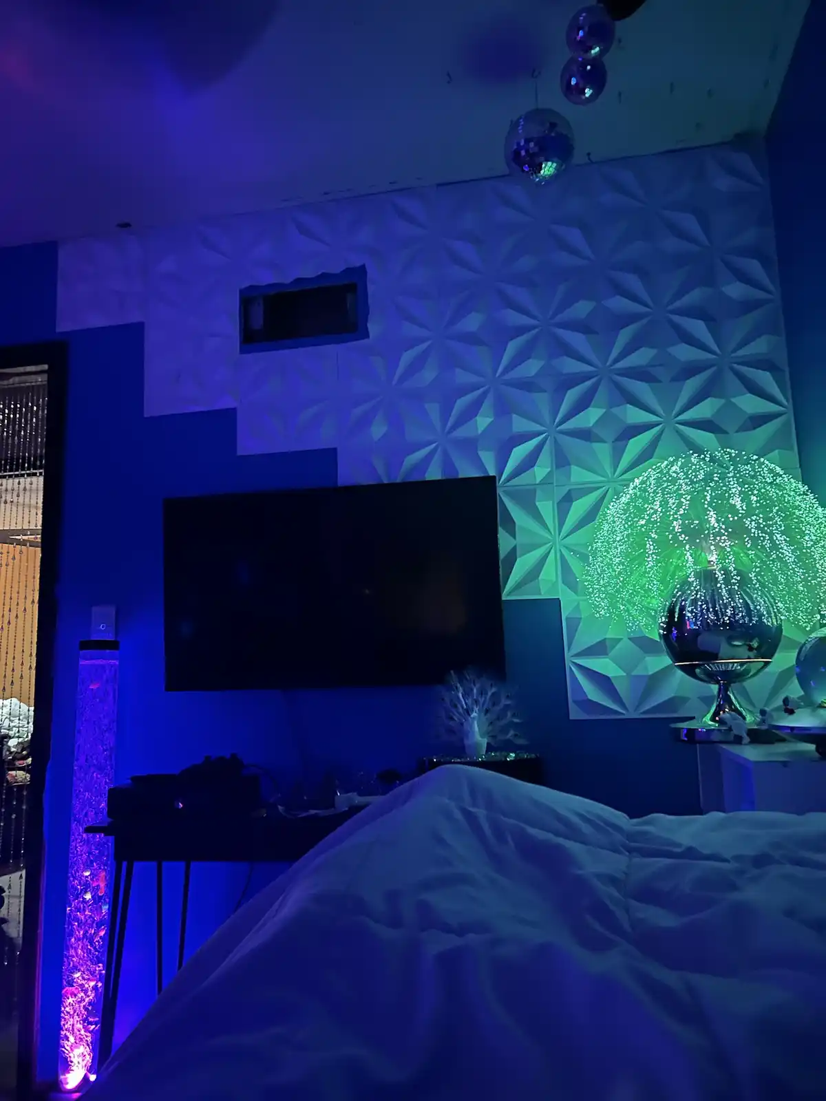 A bedroom is bathed in blue and green neon light, featuring disco balls hanging from the ceiling, a glowing green fiber…