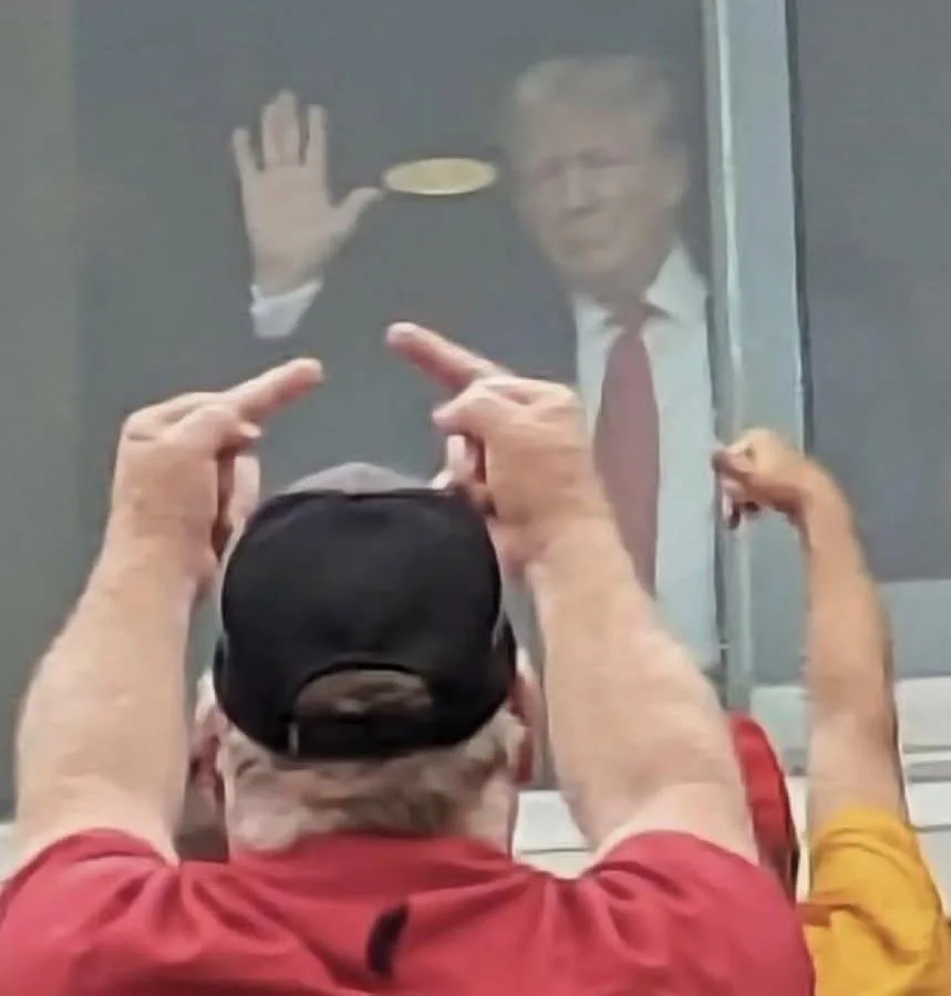 A man in a red shirt and black cap is seen from behind, pointing both index fingers at a blurry image of Donald Trump waving…