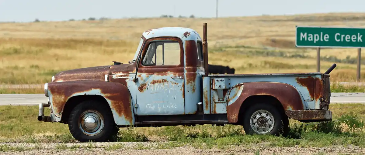 An old, rusty light blue International pickup truck sits abandoned on the side of a road in a grassy field, with a green…