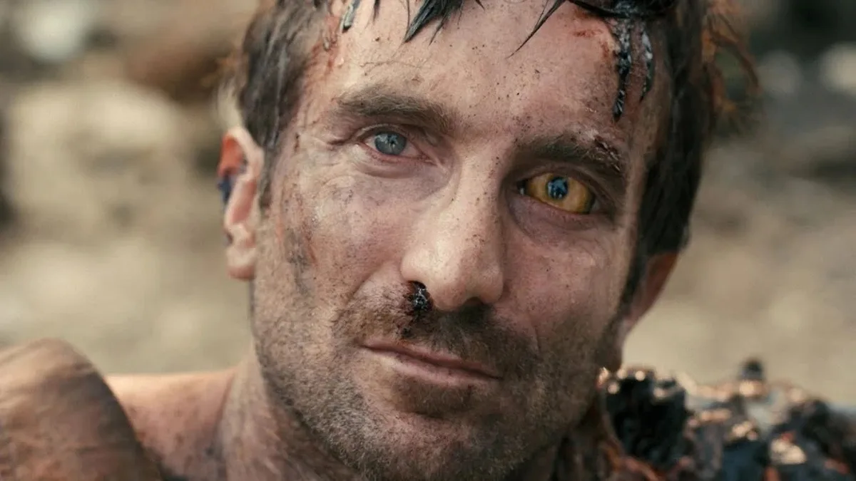 A close-up of a man with dirt and blood on his face, one eye blue and the other yellow, looking intensely at the camera. He…