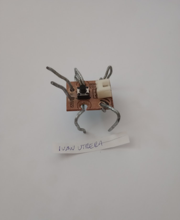 A small electronic circuit board with a push button and a white connector, modified with six bent metal wires attached to…