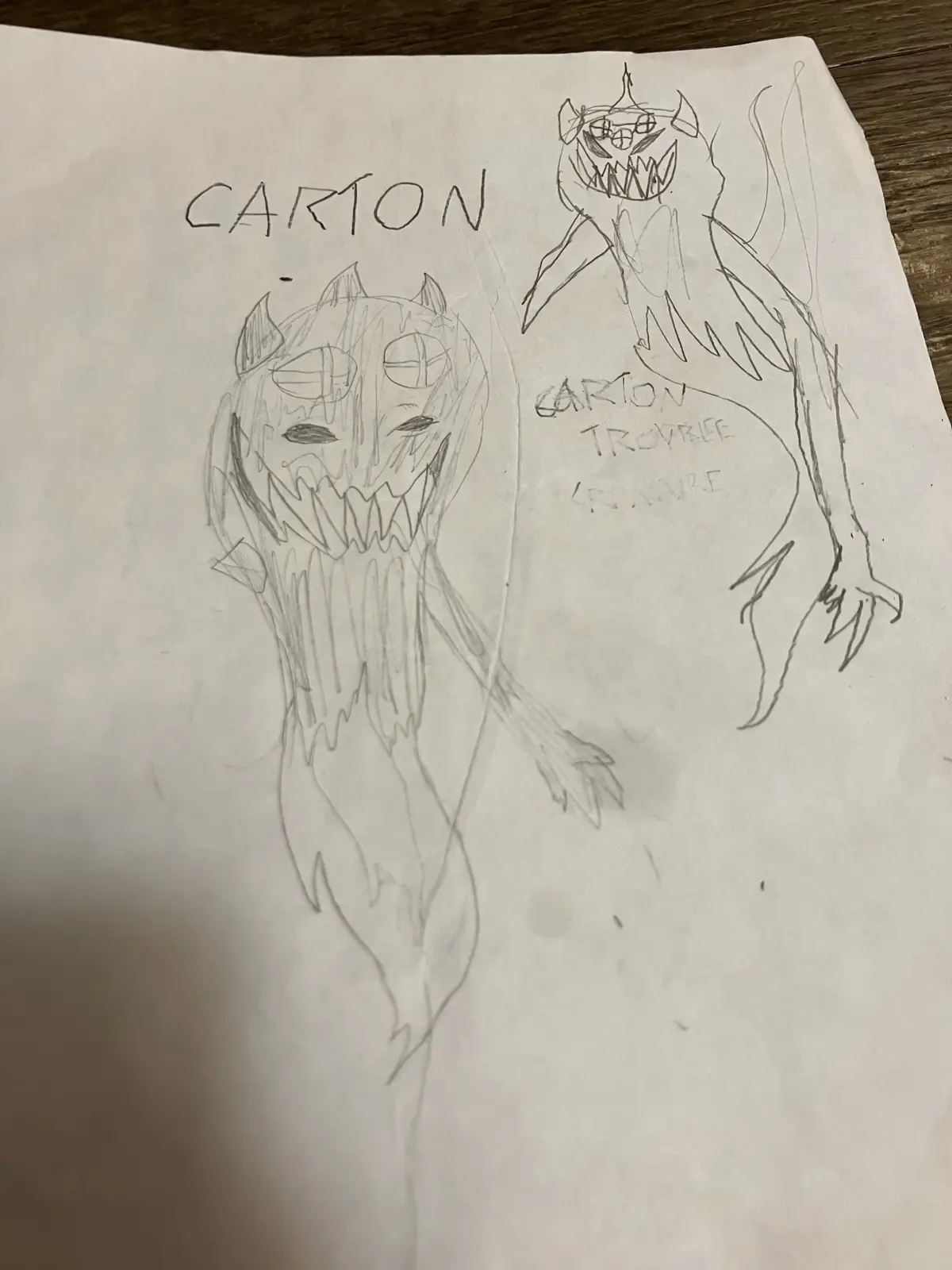 A hand-drawn pencil sketch on white paper shows two cartoonish, demonic-looking creatures with sharp teeth, horns, and…