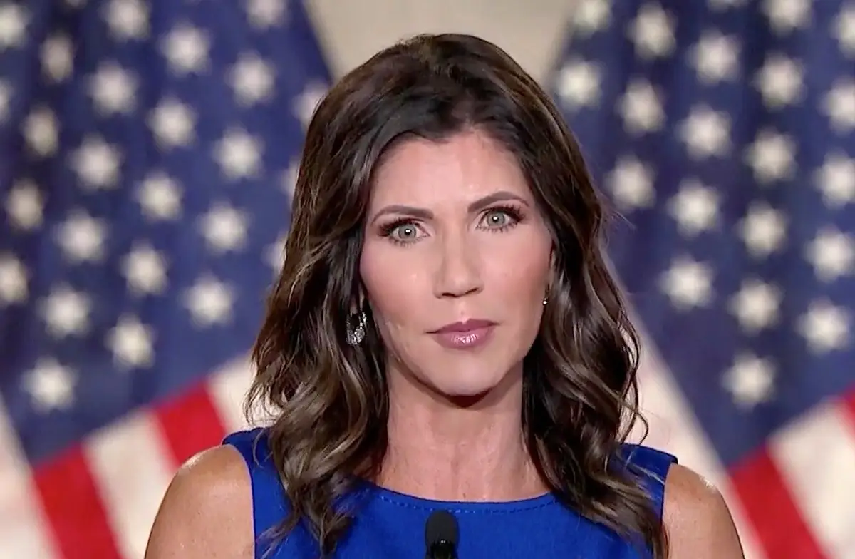 A woman with brown wavy hair and a blue dress stands in front of a blurred American flag, looking directly at the camera…