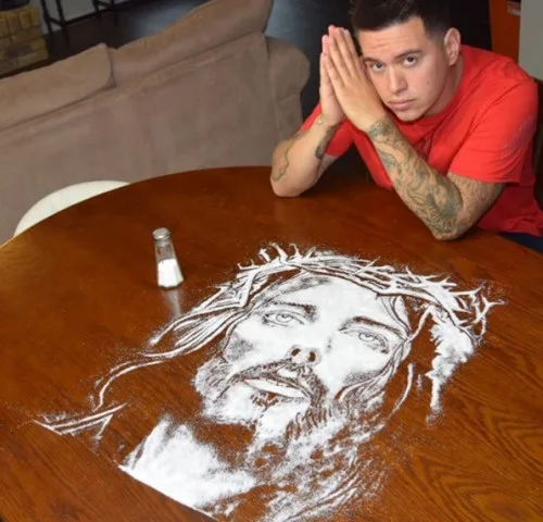 A man in a red shirt sits at a wooden table with his hands clasped near his face, looking at a detailed portrait of Jesus…