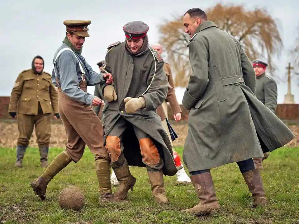 Three men in muddy World War I-era military uniforms are playing football on a grassy field; one man in the center is being…