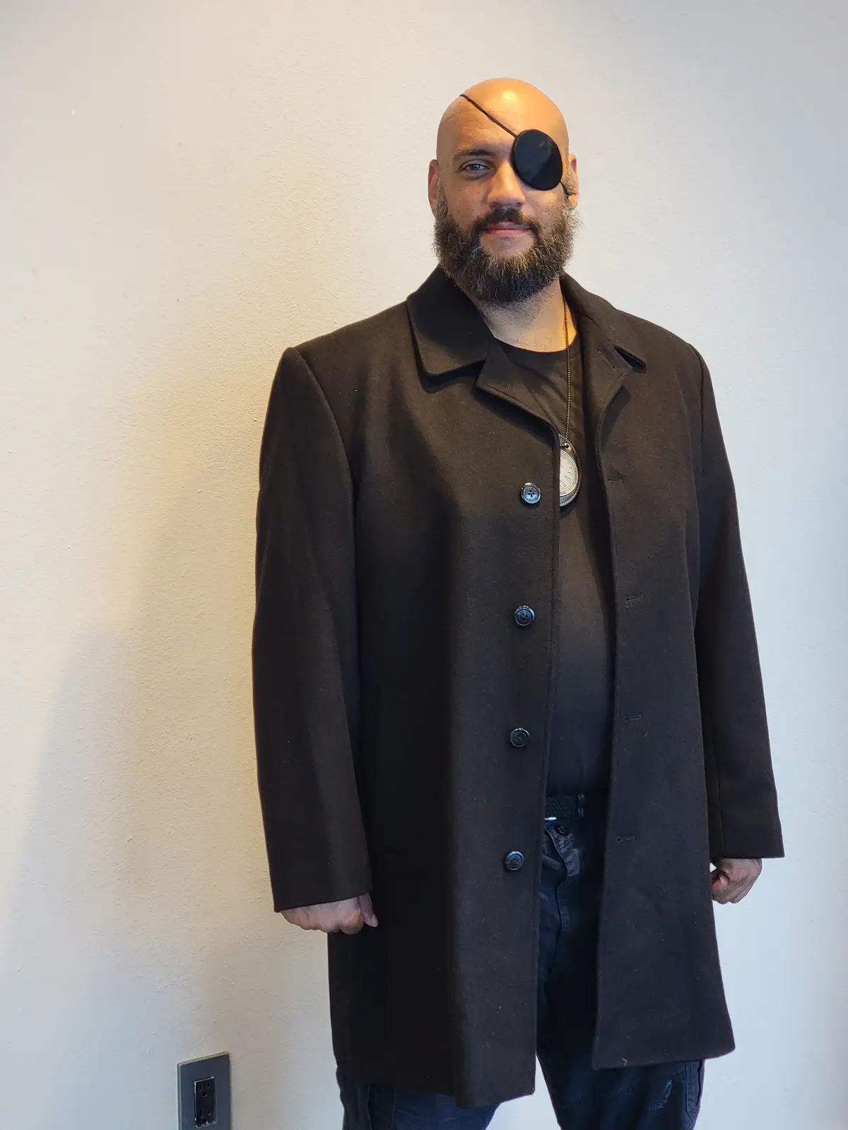 A bald man with a full beard stands against a plain wall wearing a dark coat over a t-shirt and jeans. He has a black…