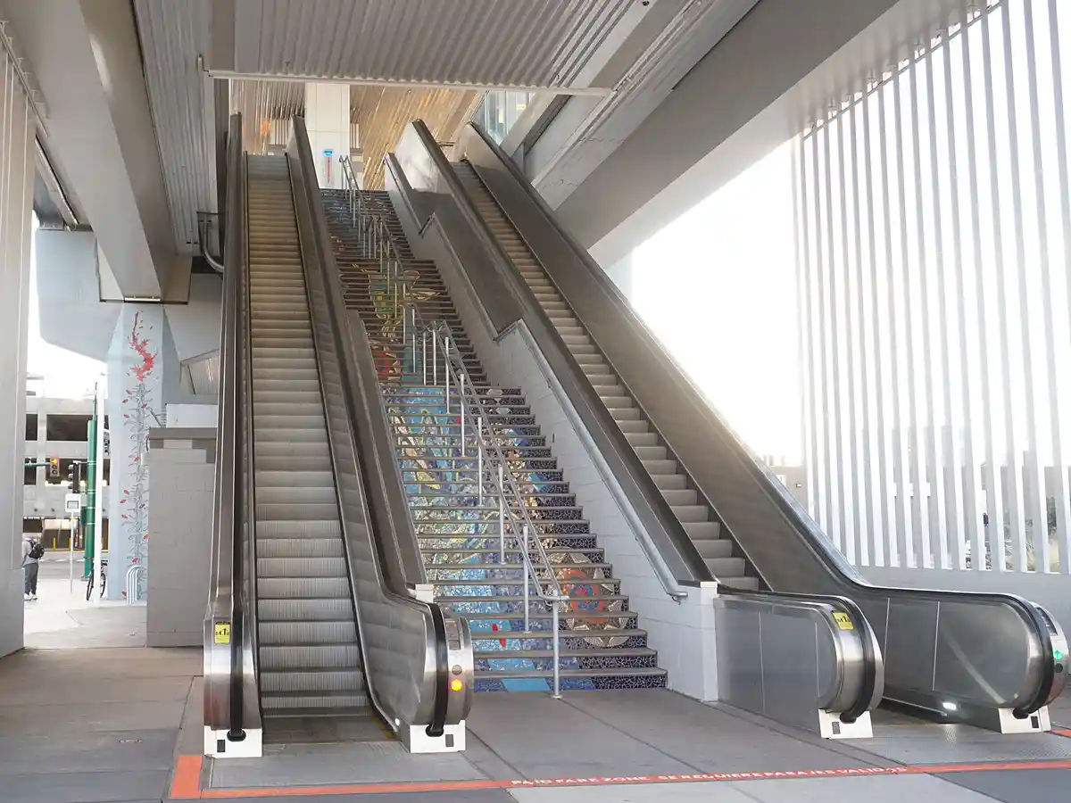 An image of a subway station entrance with two escalators flanking a central staircase decorated with colorful mosaic tiles…