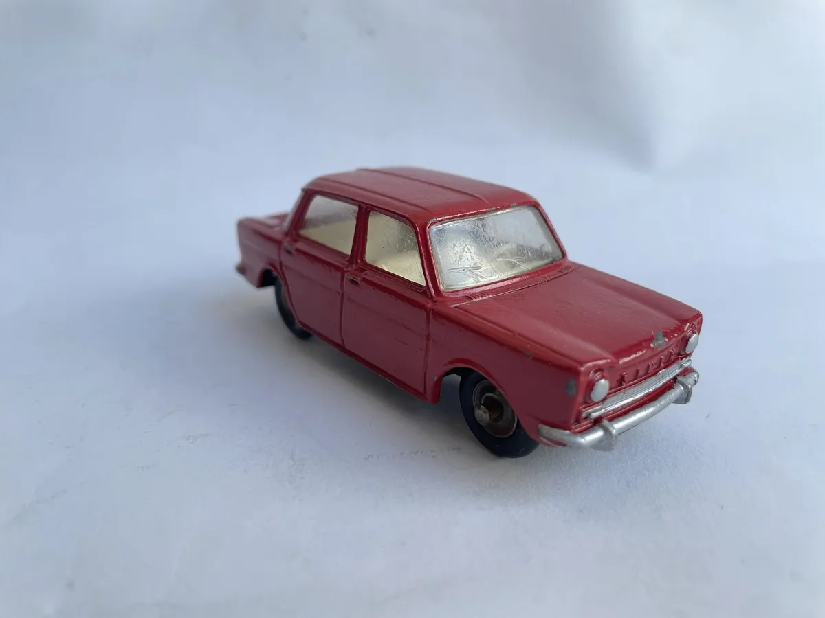 A small, red toy car with a vintage design and slightly scratched windows sits on a plain white surface. The overlaid text…