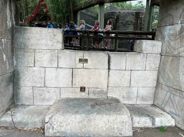 A stone wall at a zoo or wildlife park features two small plaques, one reading 'this is not a drill' and the other 'this is…