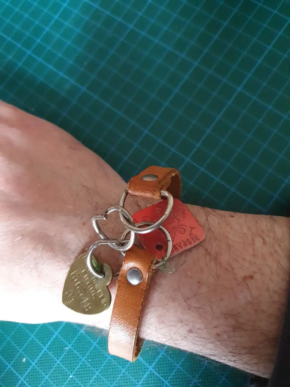 A person's wrist is shown wearing a brown leather bracelet that has been modified into a keychain, with a red dog tag and a…