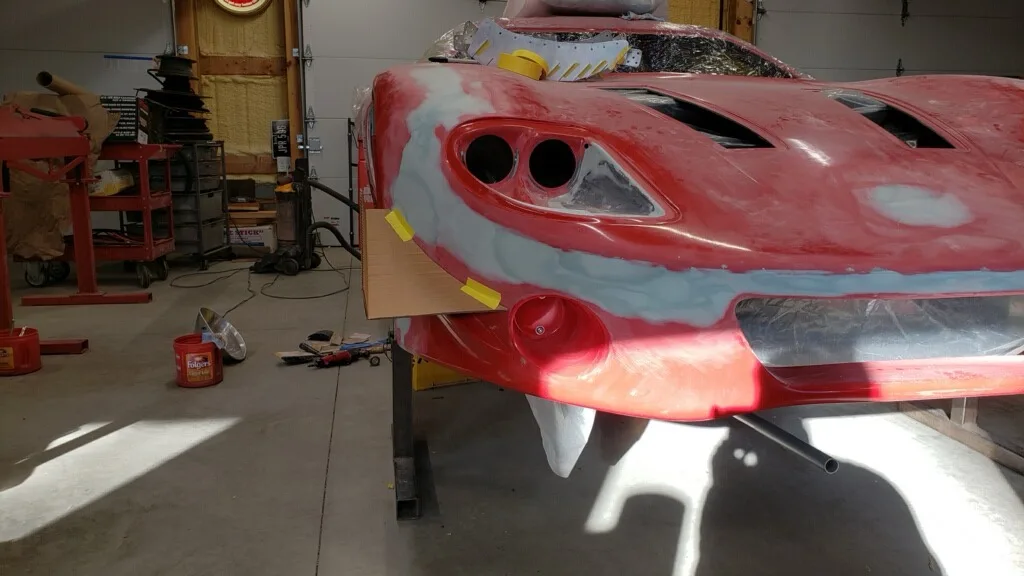 A red sports car body is shown in a garage, partially painted with white primer or filler, with visible holes and rough…