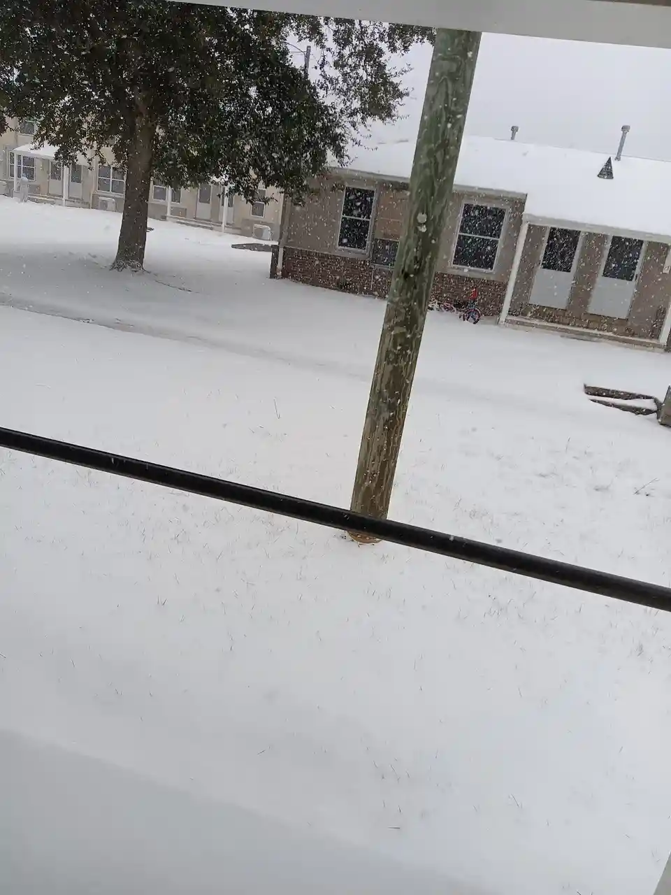 A snowy suburban street viewed from a porch, with snow-covered lawns, houses, and trees, and a child's bike visible near a…
