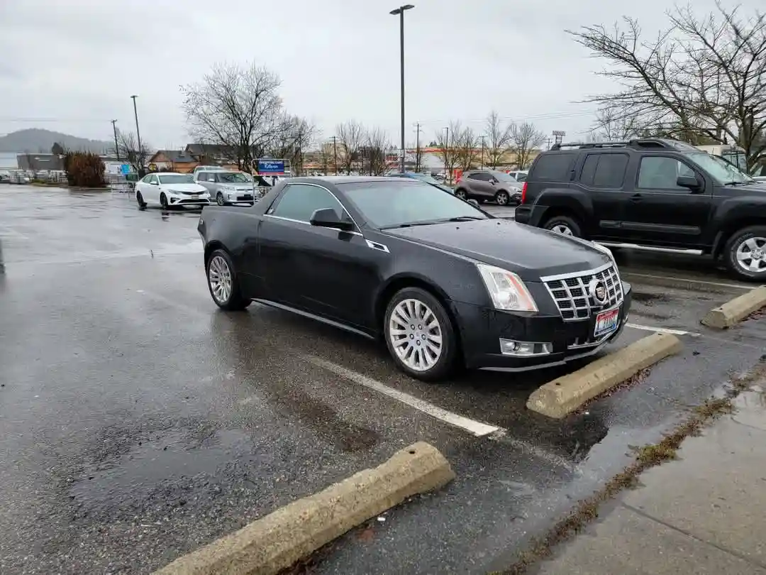 A black Cadillac convertible is parked in a wet parking lot, angled slightly toward the viewer, with its top down. The car…