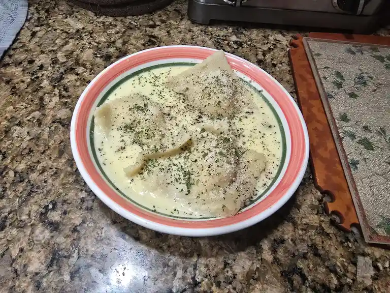 A bowl of ravioli in a creamy sauce, topped with grated cheese and dried herbs, sits on a speckled granite countertop. The…