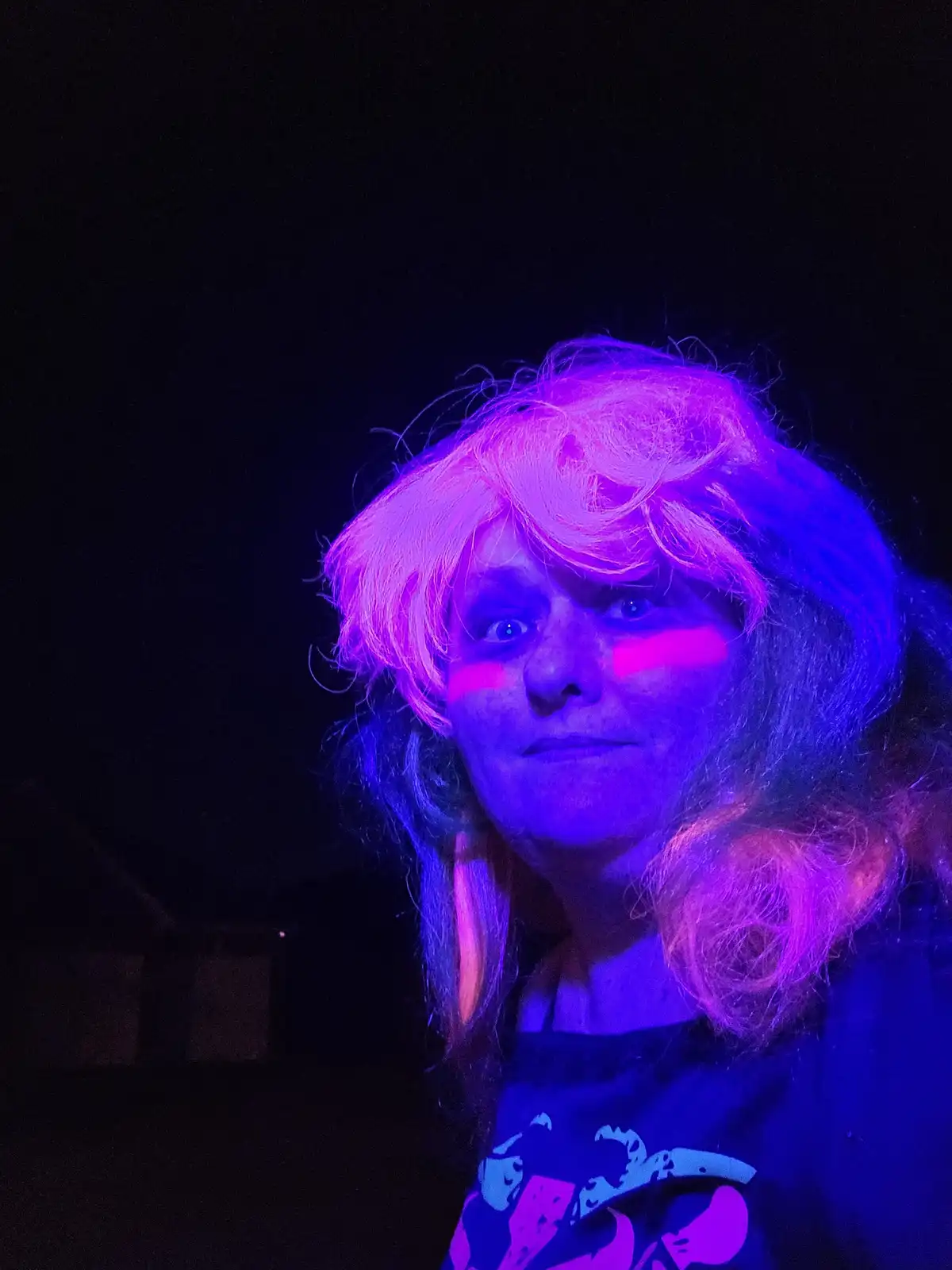 A person with a long, wavy wig under intense purple and pink lighting, with two bright pink horizontal stripes painted on…