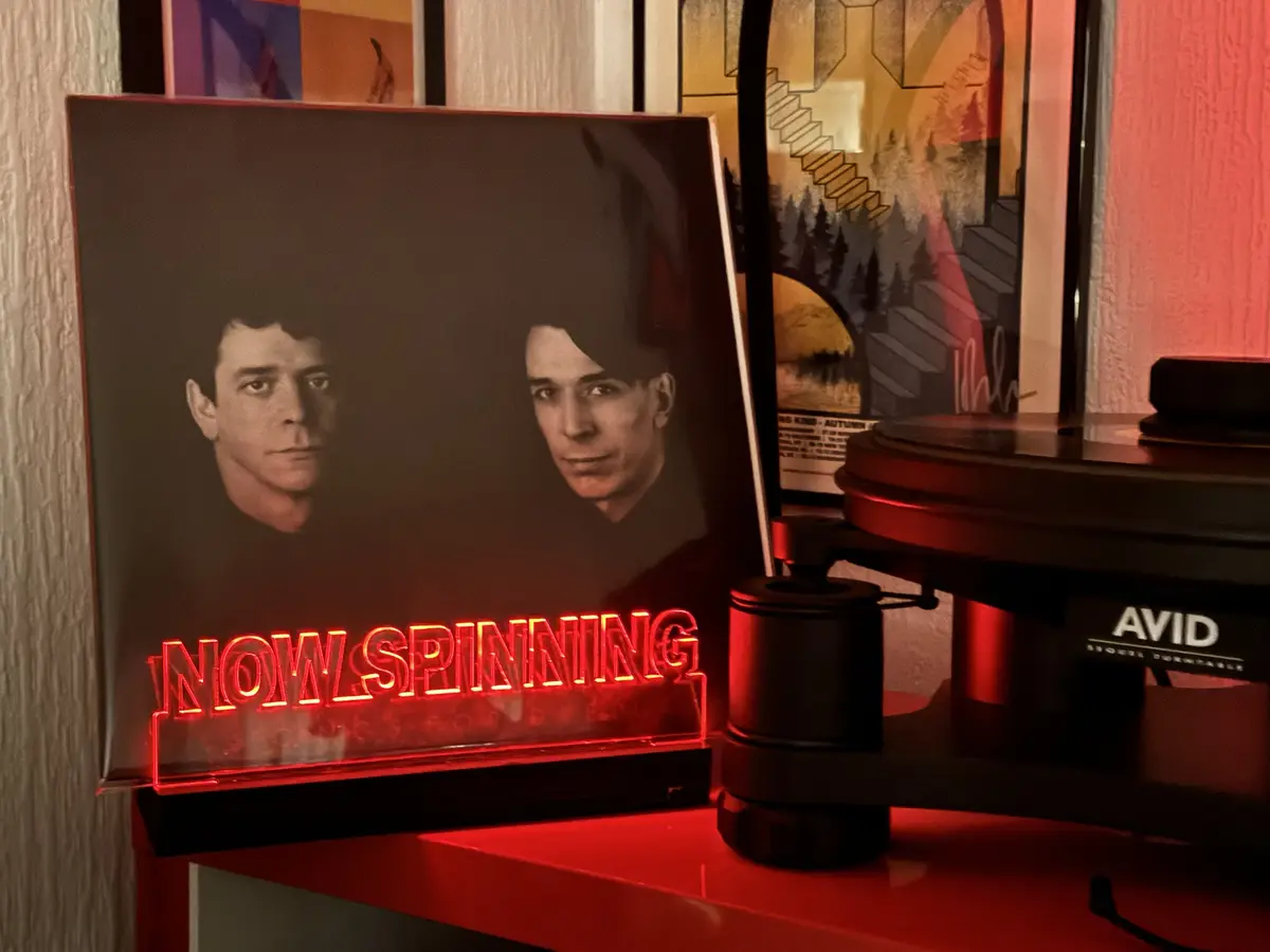 A vinyl record sleeve featuring two men is propped up next to an AVID turntable, with a red neon sign below it that reads…