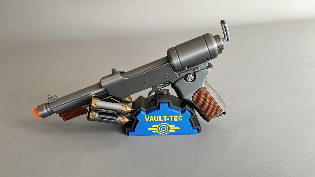 A silver toy gun with orange tip and brown grips rests on a blue and black stand featuring the yellow 'VAULT-TEC' logo and…