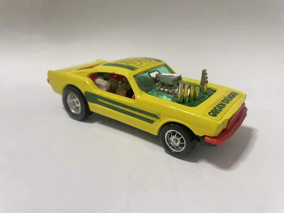A yellow toy car with green stripes and a visible engine, has the text 'ORGAN GRINDER' written on its rear. The car's engine…