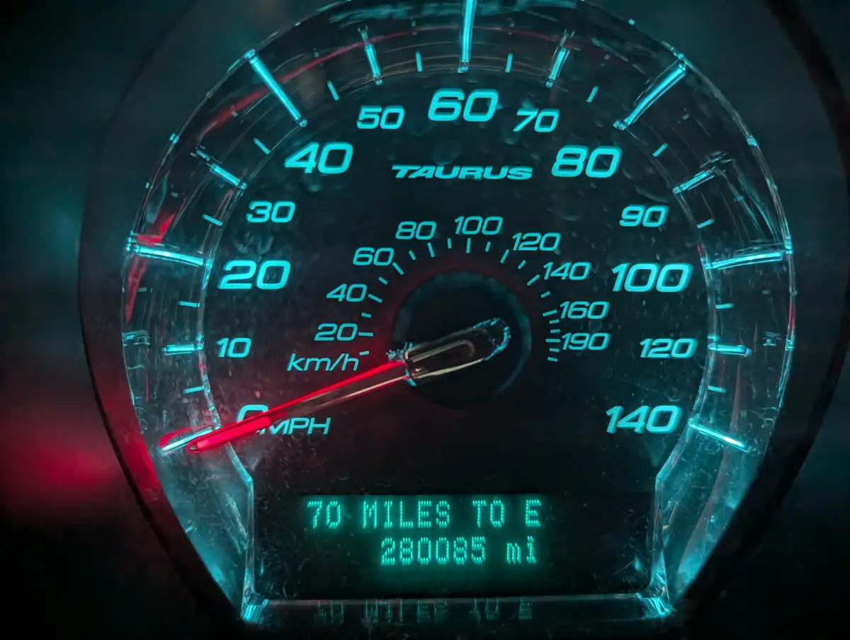 A close-up of a Ford Taurus speedometer with glowing blue markings and a red needle at zero, showing a digital odometer…