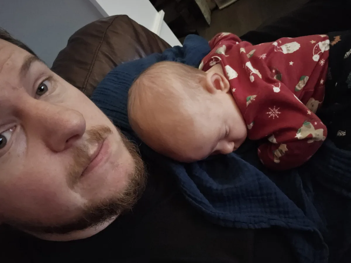 A man with a mustache and beard looks at the camera with a tired but loving expression while holding a sleeping baby dressed…