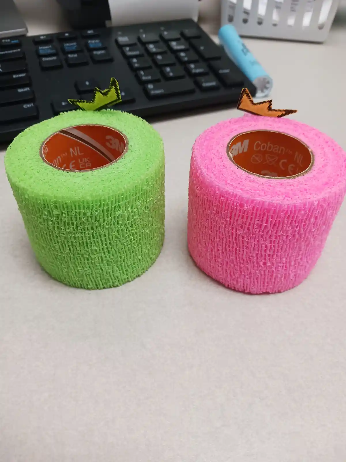 Two rolls of 3M Coban medical elastic bandage, one green and one pink, are arranged on a desk to resemble apples, each…