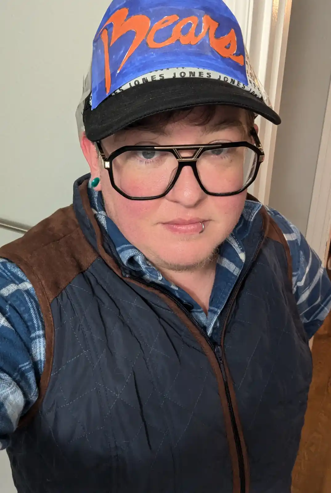 A person wearing oversized black-rimmed glasses, a blue plaid shirt under a dark quilted vest, and a baseball cap with…