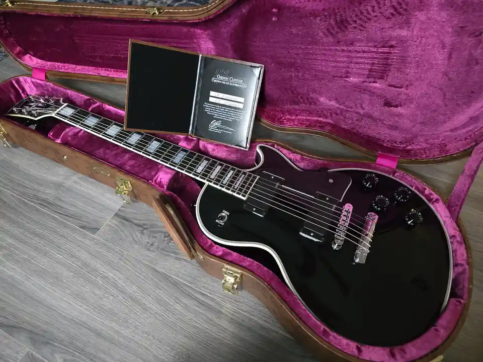 A black Gibson Custom electric guitar rests in an open case with magenta velvet lining, next to its official certificate of…
