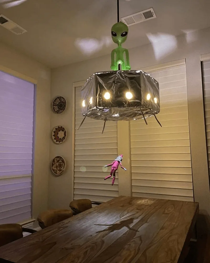 A green alien figurine sits atop a homemade chandelier wrapped in foil, with a pink elf doll floating mid-air nearby. The…